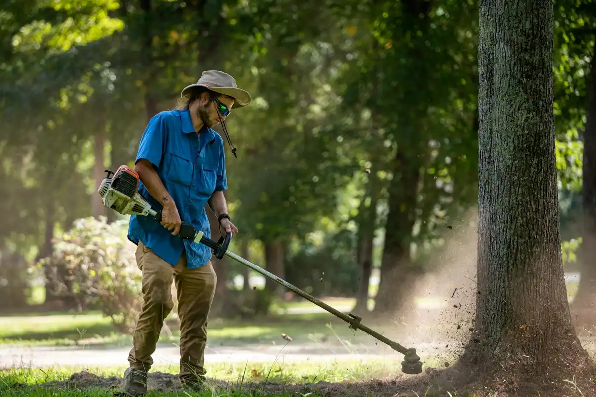 A person wearing a blue shirt, khaki pants, and a wide-brimmed hat mows the lawn with a string trimmer beside a tree in a sunlit, park-like yard.