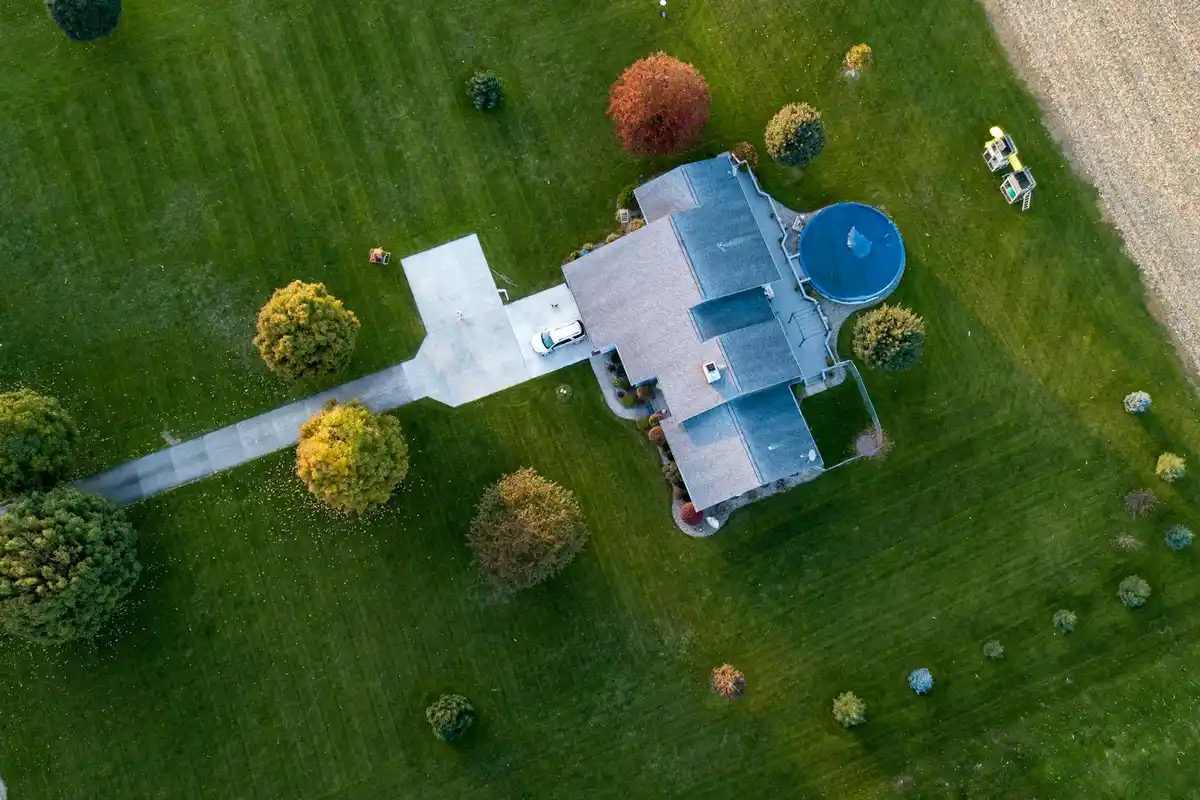 Aerial view of a house surrounded by a large, manicured green lawn with scattered trees and a circular pool.