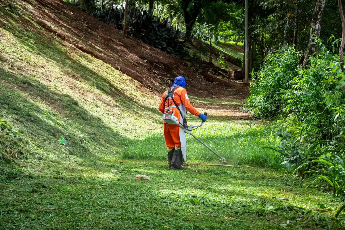 Lawn maintenance worker in orange safety gear using a brush cutter on a sloped, grassy hillside.