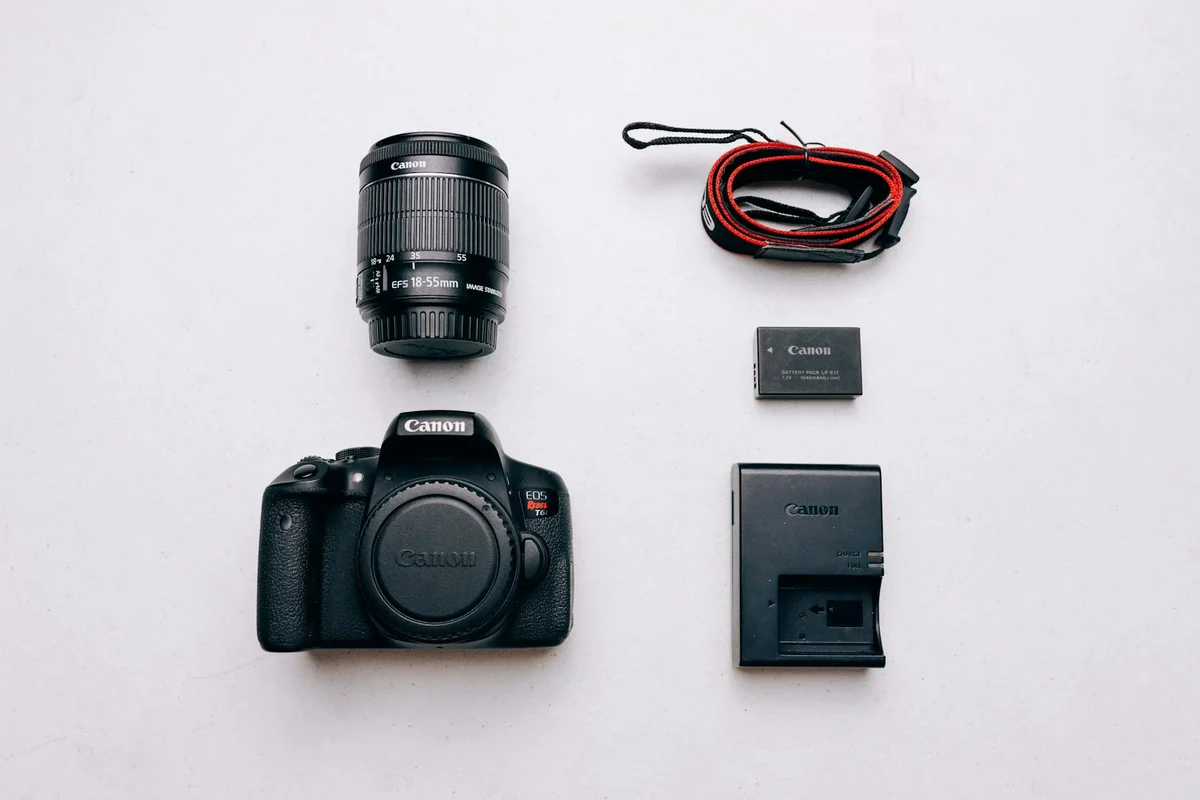 Canon DSLR camera with a zoom lens, spare battery, and battery charger arranged on a light background