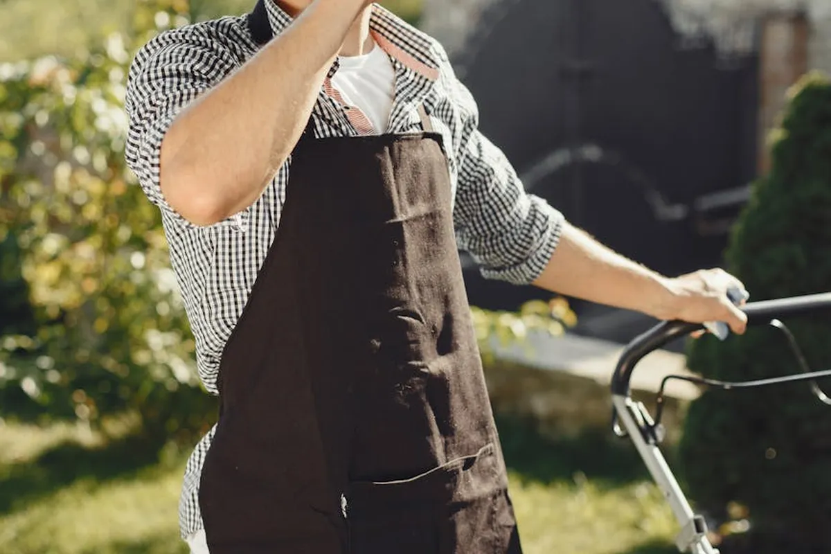 Gardener wearing a checkered shirt and brown apron stands beside a lawn mower in a sunny yet shady backyard.