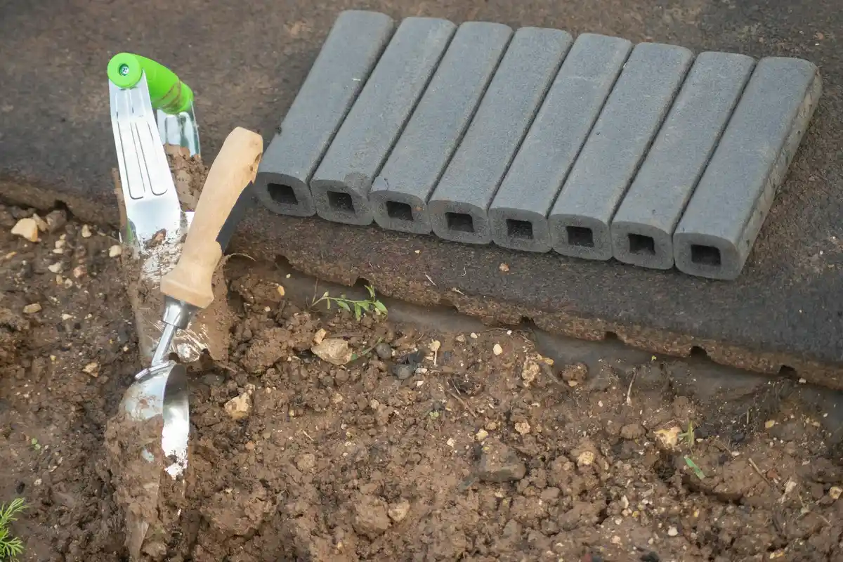 Long-handled garden aeration tool on bare soil beside a row of hollow concrete blocks.