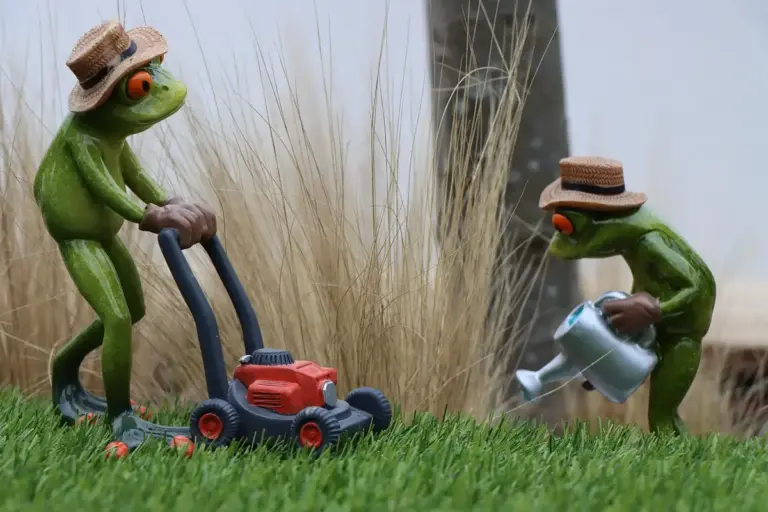 Two green frog figurines wearing straw hats in a yard; one is pushing a red lawn mower while the other waters the grass with a small silver watering can.