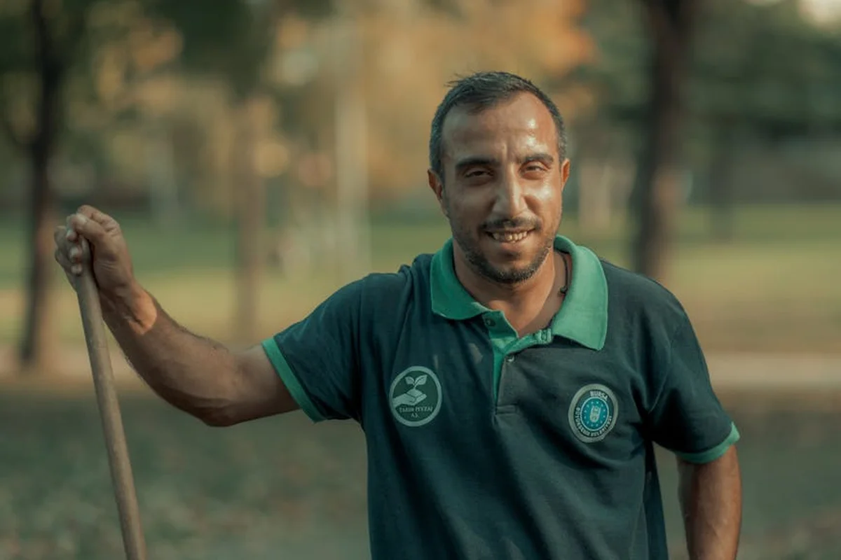 A man in a green polo shirt holds a rake in a park, preparing for lawn maintenance.