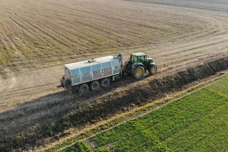 Aerial view of a tractor pulling a large fertilizer spreader along a field, with bare soil on one side and green crops on the other.