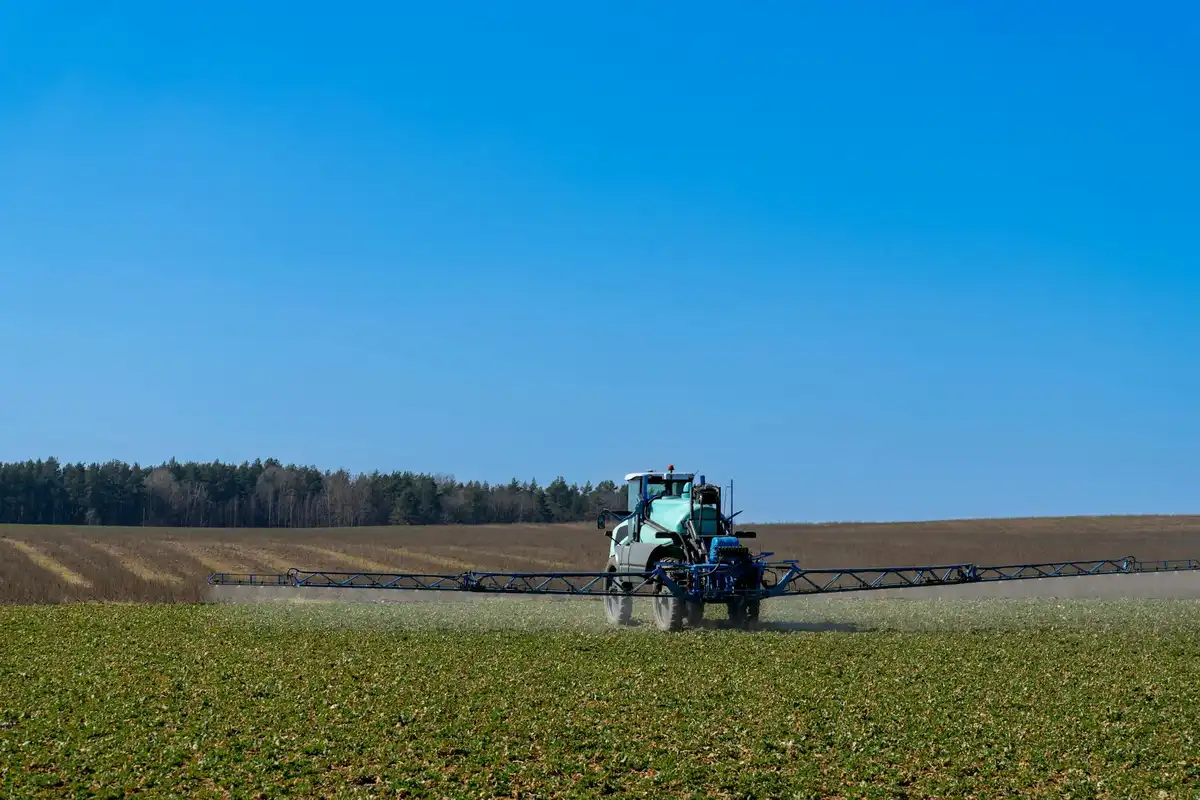 Green tractor with a wide-area fertilizer spreader working across a field under a clear blue sky.