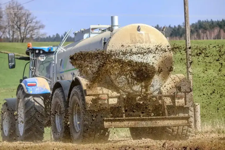 Blue tractor pulling a beige liquid fertilizer tanker spreading material across a field.