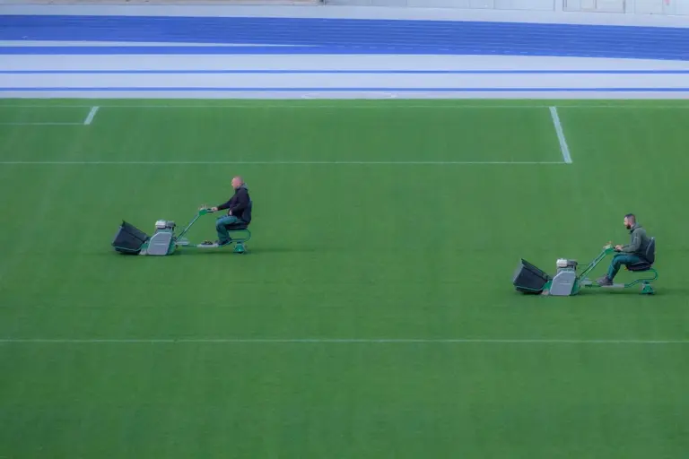 Two people riding lawn mowers mowing a large green sports field.