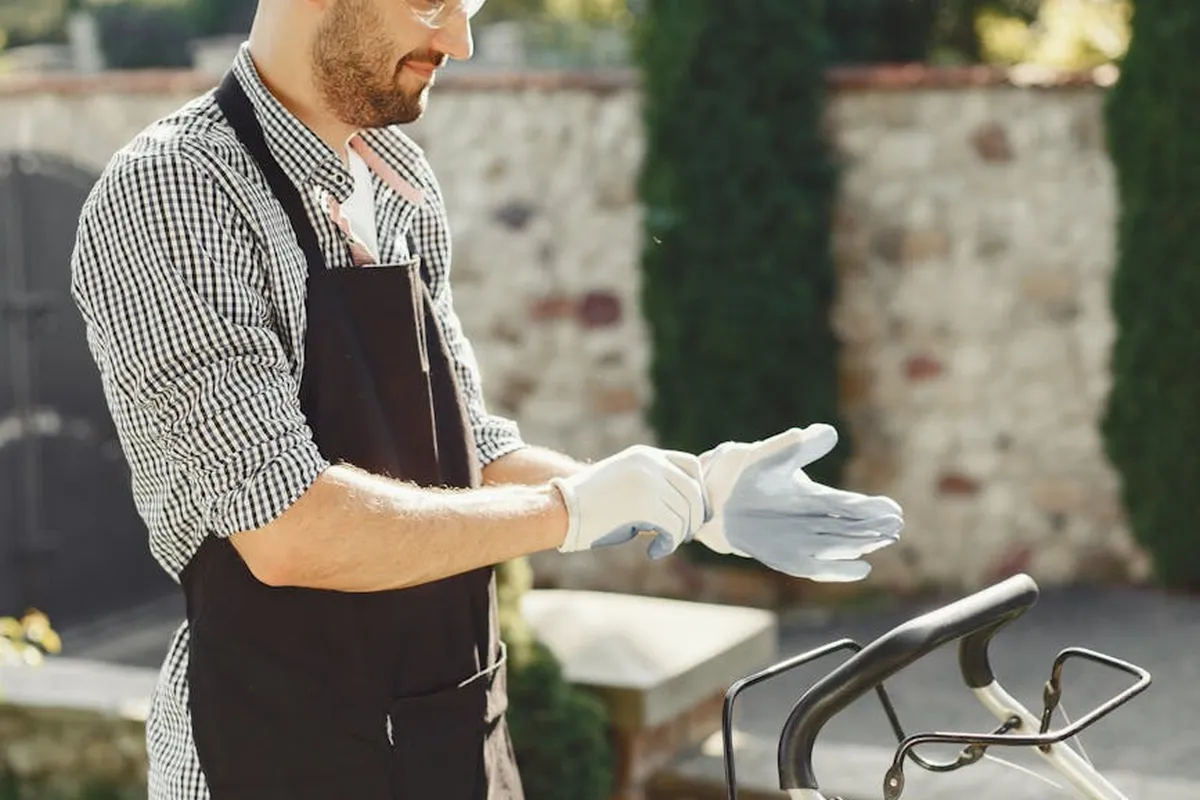 A man wearing a checkered shirt, gloves, and an apron stands beside a lawn mower outdoors, preparing to start it.