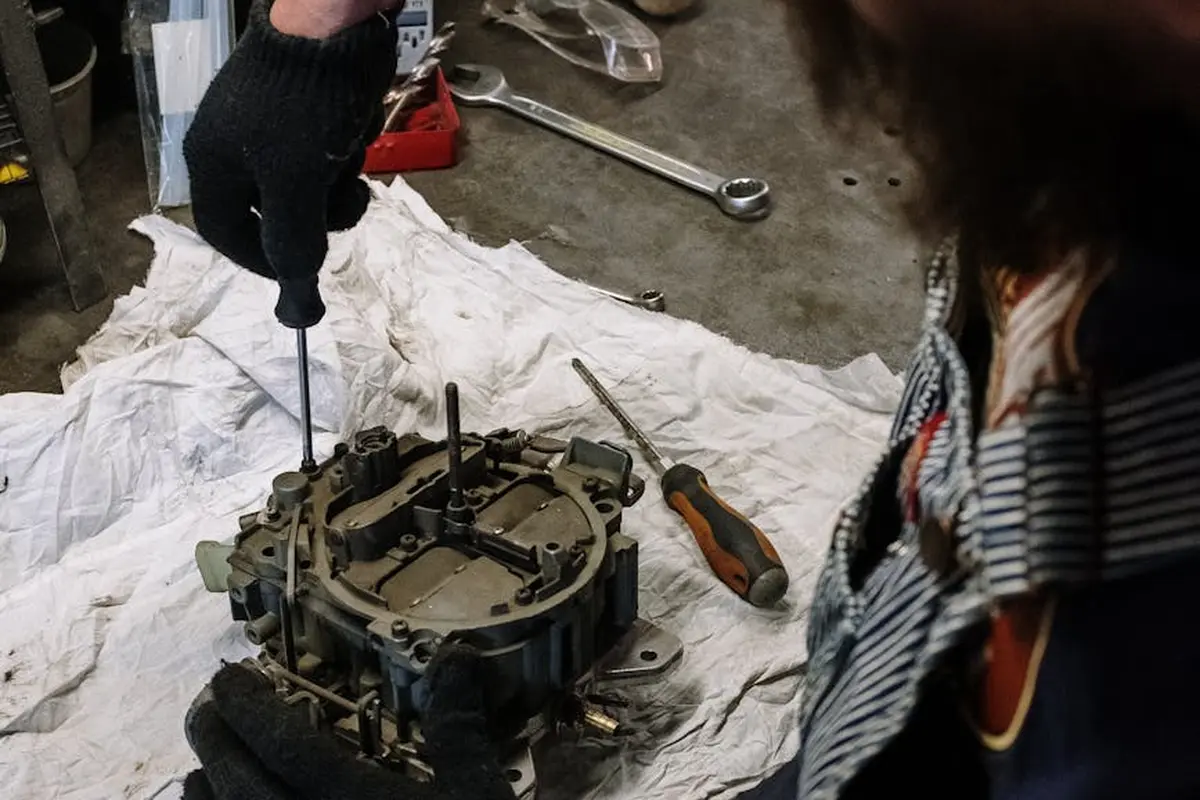 Person wearing gloves examining a carburetor assembly on a white cloth, with tools in the background.