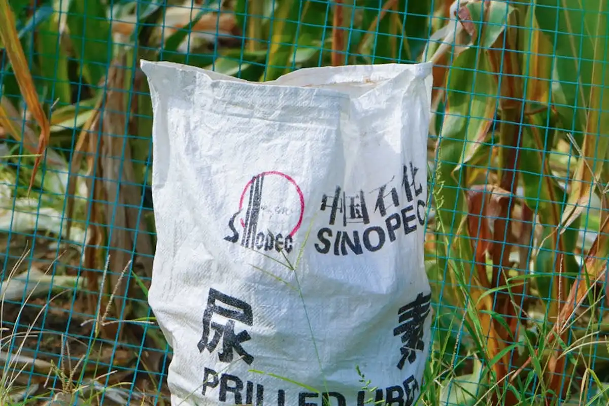 White bag of fertilizer with Chinese characters and a red logo, placed behind green garden netting.