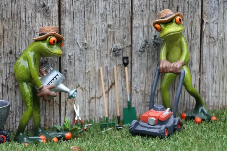 Two green frogs wearing hats stand among garden tools and a toy lawn mower in a grassy yard