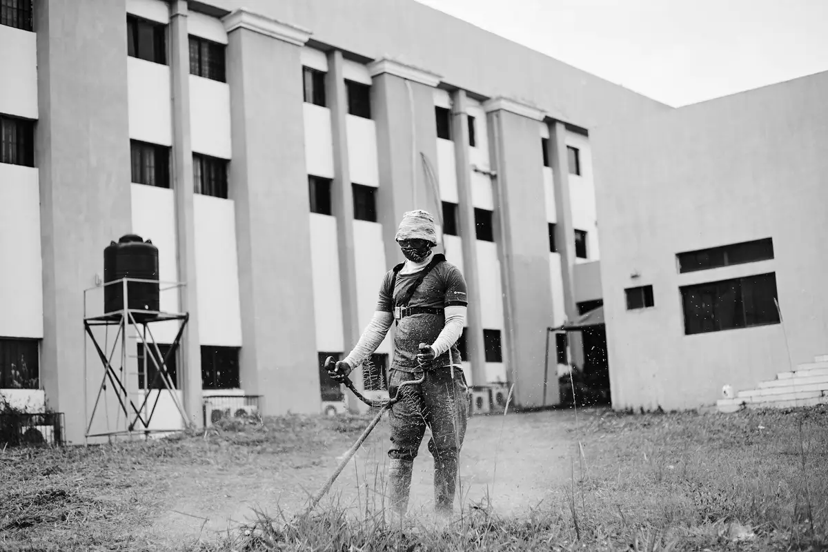 Person wearing protective gear and a mask standing in a grassy area beside a large building, holding a gasoline-powered lawn tool.