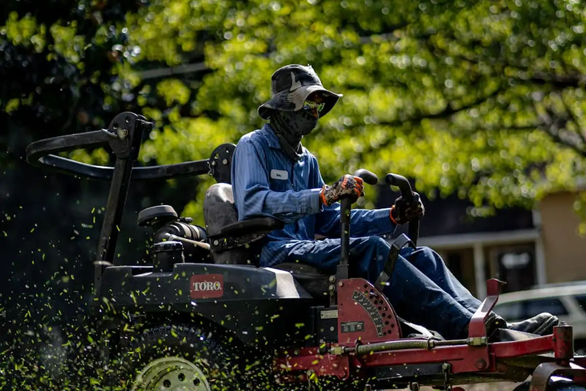 A person in protective clothing sits on a riding lawn mower, cutting grass with green foliage in the background.