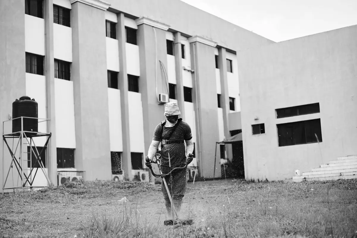 Black-and-white photo of a person wearing protective gear and a mask, tending a lawn in front of a modern building.