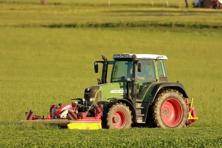 Green lawn tractor with red wheels operating a mower attachment in a sunlit grassy field.