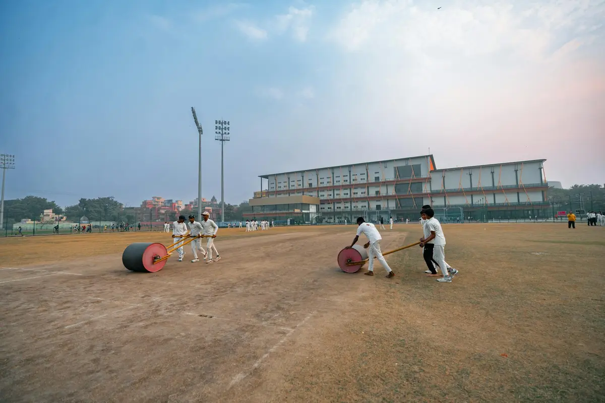 A group of people pushing a large lawn roller across a dusty sports field with a building in the background.