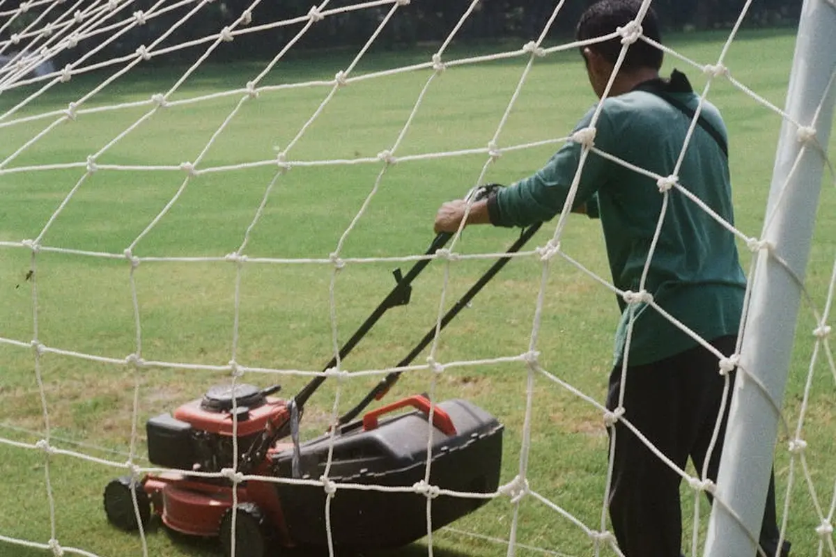 A person wearing a green shirt pushes a lawn mower along a grassy field behind a soccer goal, illustrating post-aeration lawn care.
