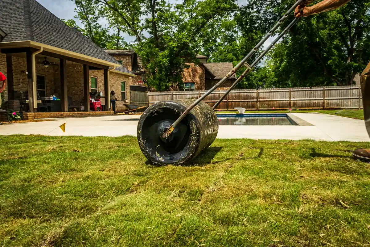 Close-up of a lawn aerator wheel on a green backyard lawn with a house, pool, and wooden fence in the background