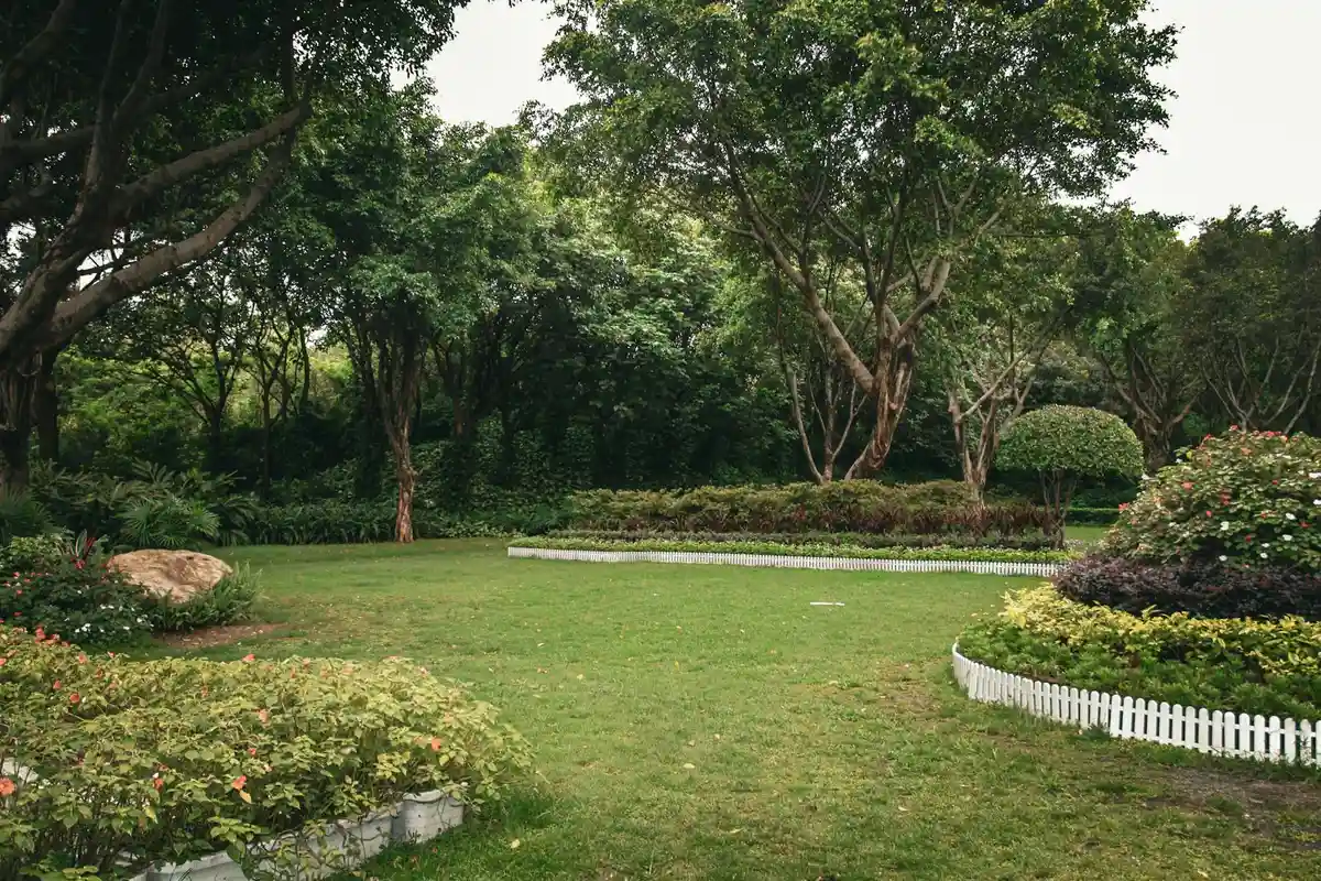 A well-kept lawn with lush green grass, mature trees, and flowerbeds edged by a white fence.