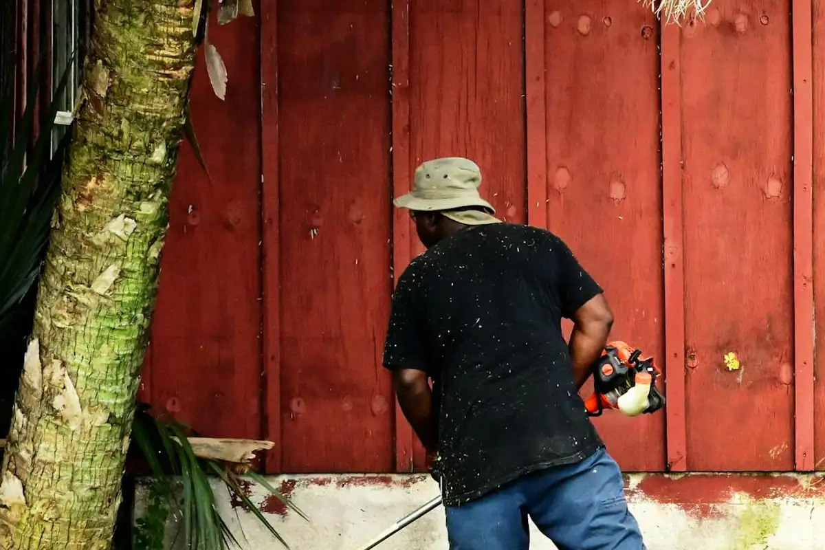 A person wearing a wide-brimmed hat and dark shirt uses a lawn tool near a red wooden fence