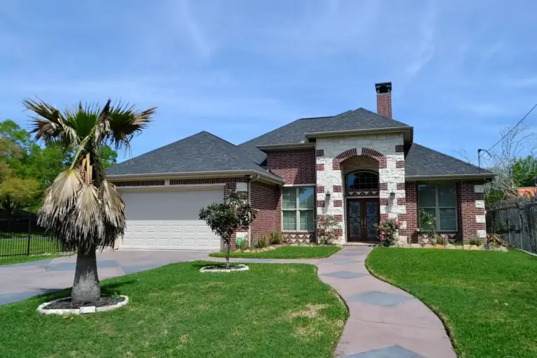 Front yard with a curved concrete path leading to a brick house, green lawn, and a small palm tree on the left under a clear blue sky.