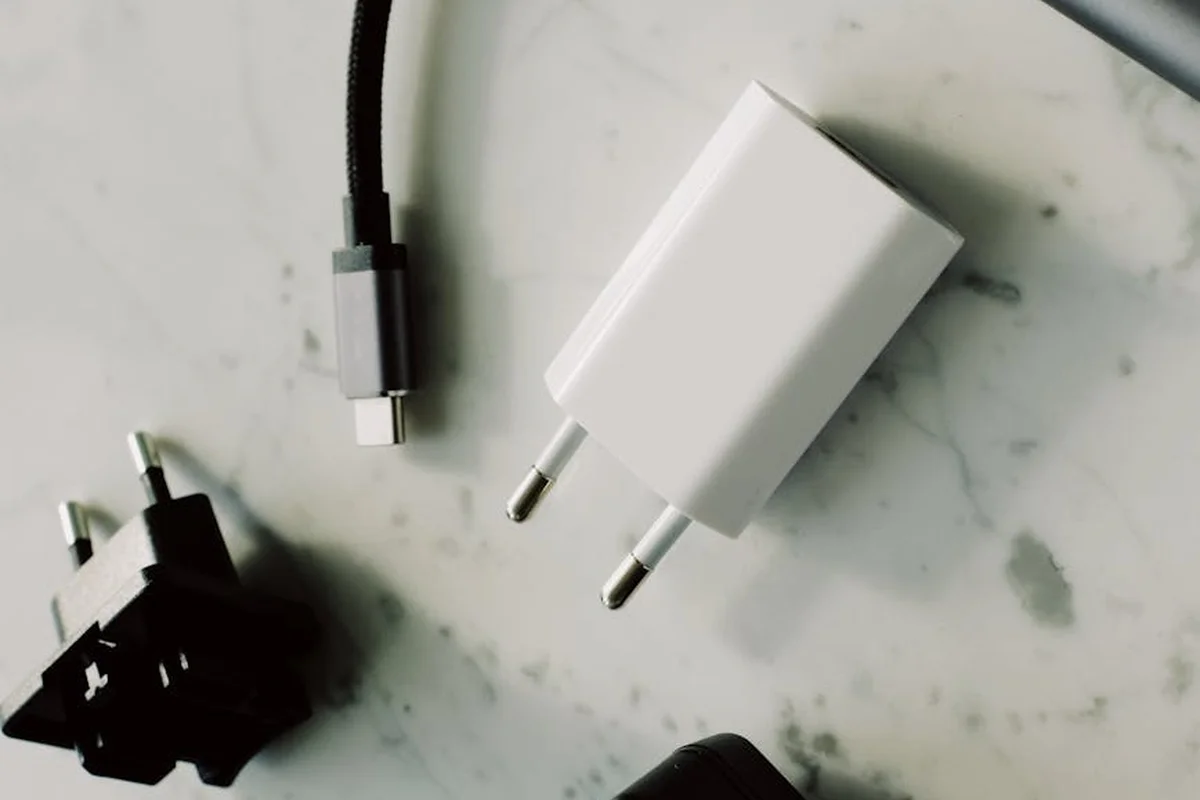 Close-up of electrical charging components on a light surface: a white wall charger, a USB-C cable, and connectors, representing lawn mower battery charging gear.