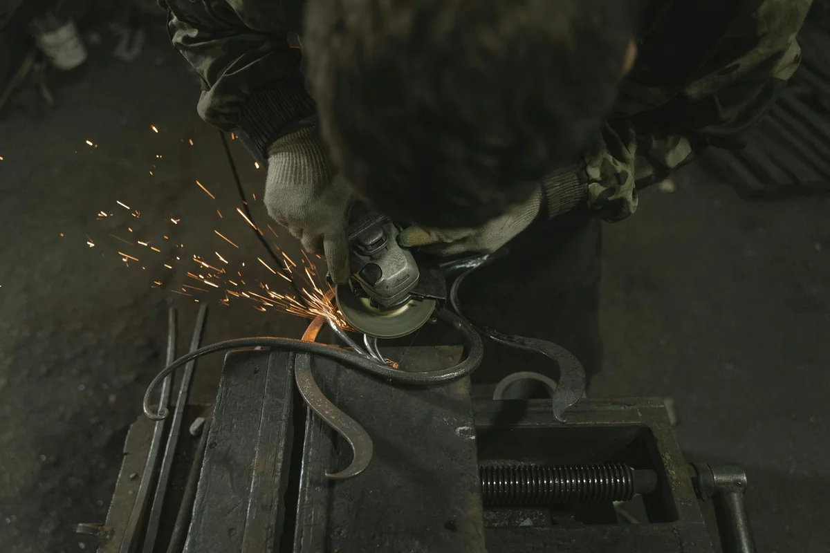Worker wearing protective gear sharpens a lawn mower blade with a grinder, sparks flying as metal chips off the blade.