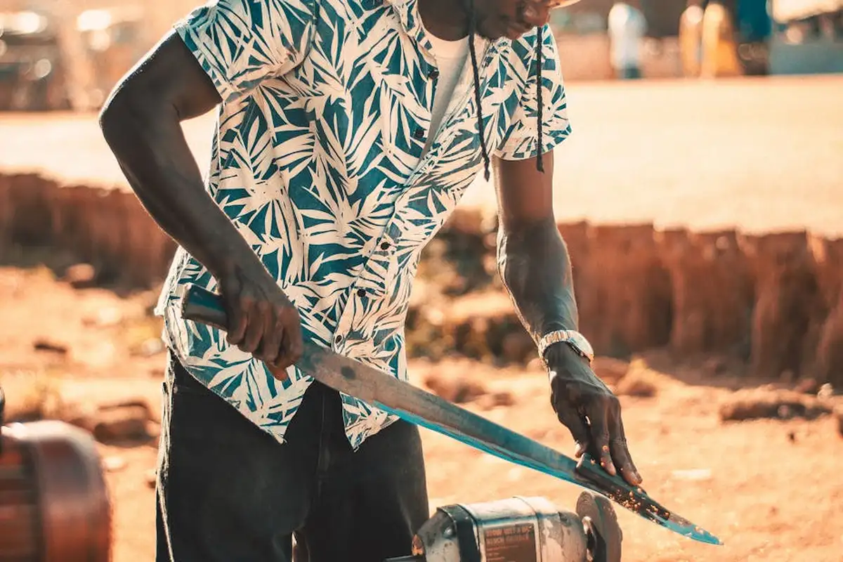 Person in a tropical-pattern shirt sharpening a lawn mower blade with a grinder outdoors.