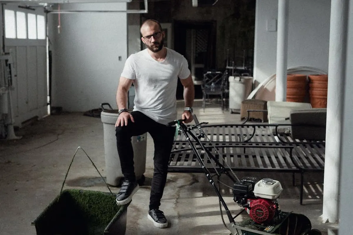 Bearded man in a white t-shirt and dark jeans stands beside a gas-powered lawn mower in a garage-like workshop.