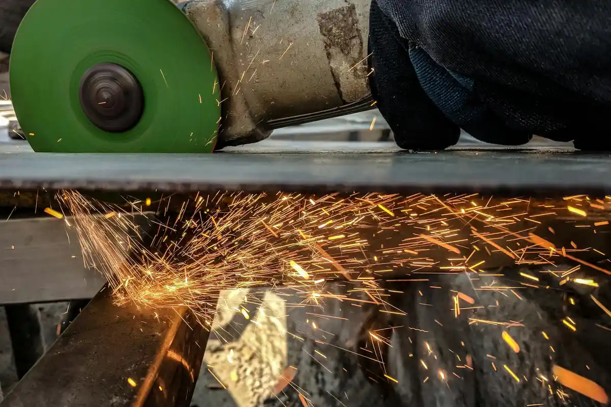 Close-up of a mower blade being sharpened on a green grinding wheel, with sparks flying and a gloved hand steadying the metal.