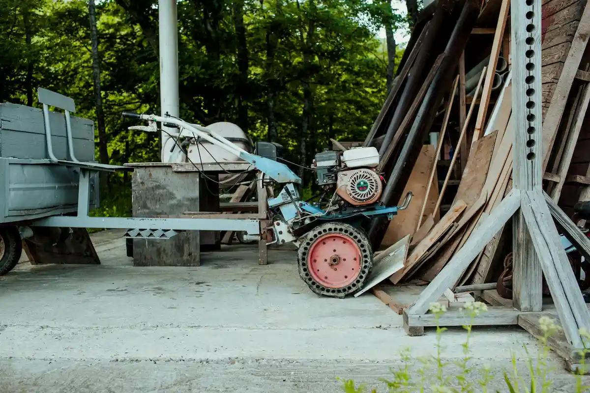 Power lawn tool (tiller/mower) on a concrete surface, with a trailer on the left and a stack of lumber on the right.
