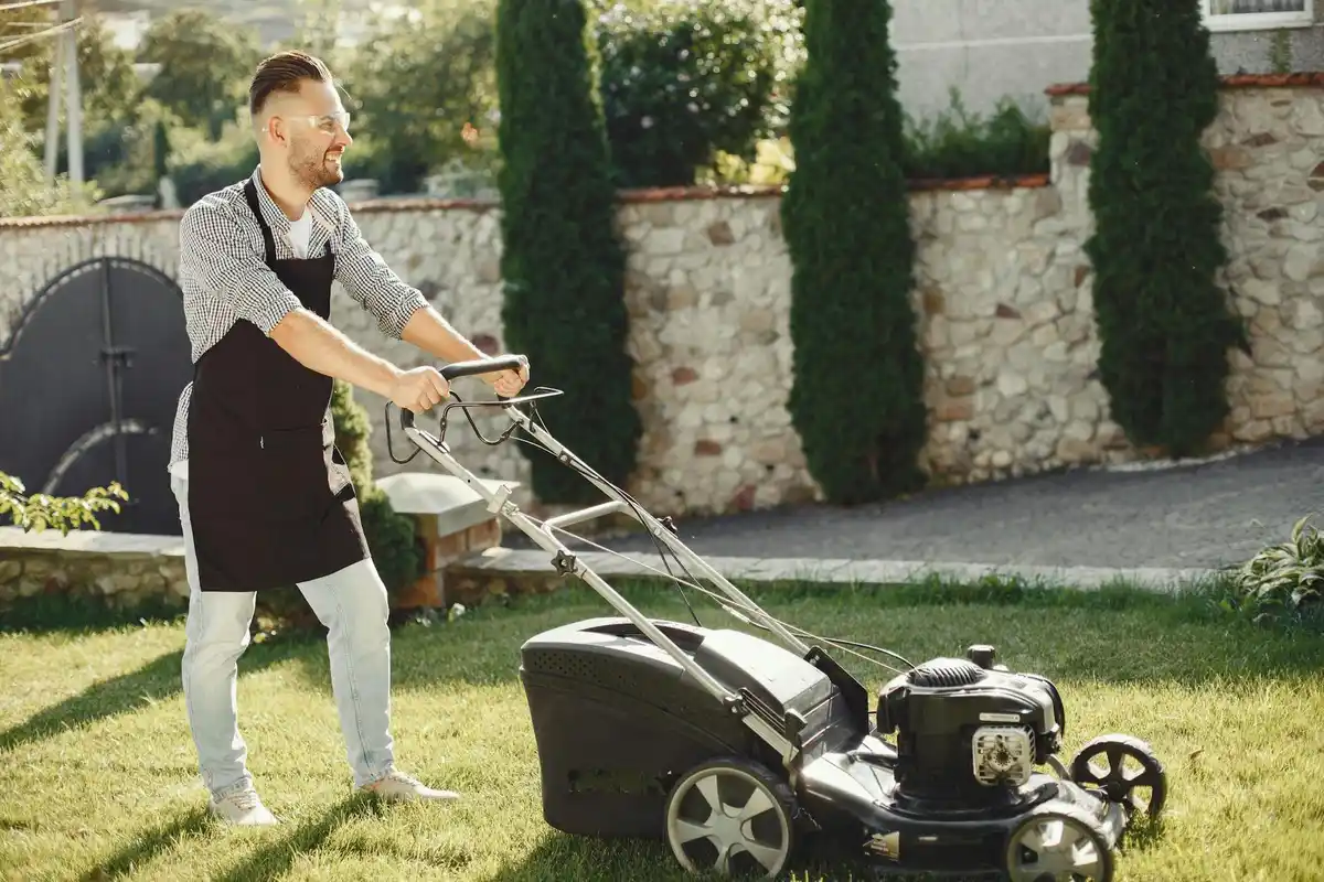 A man wearing casual clothing pushes a lawn mower across a sunlit residential lawn with a stone wall and evergreen trees in the background.