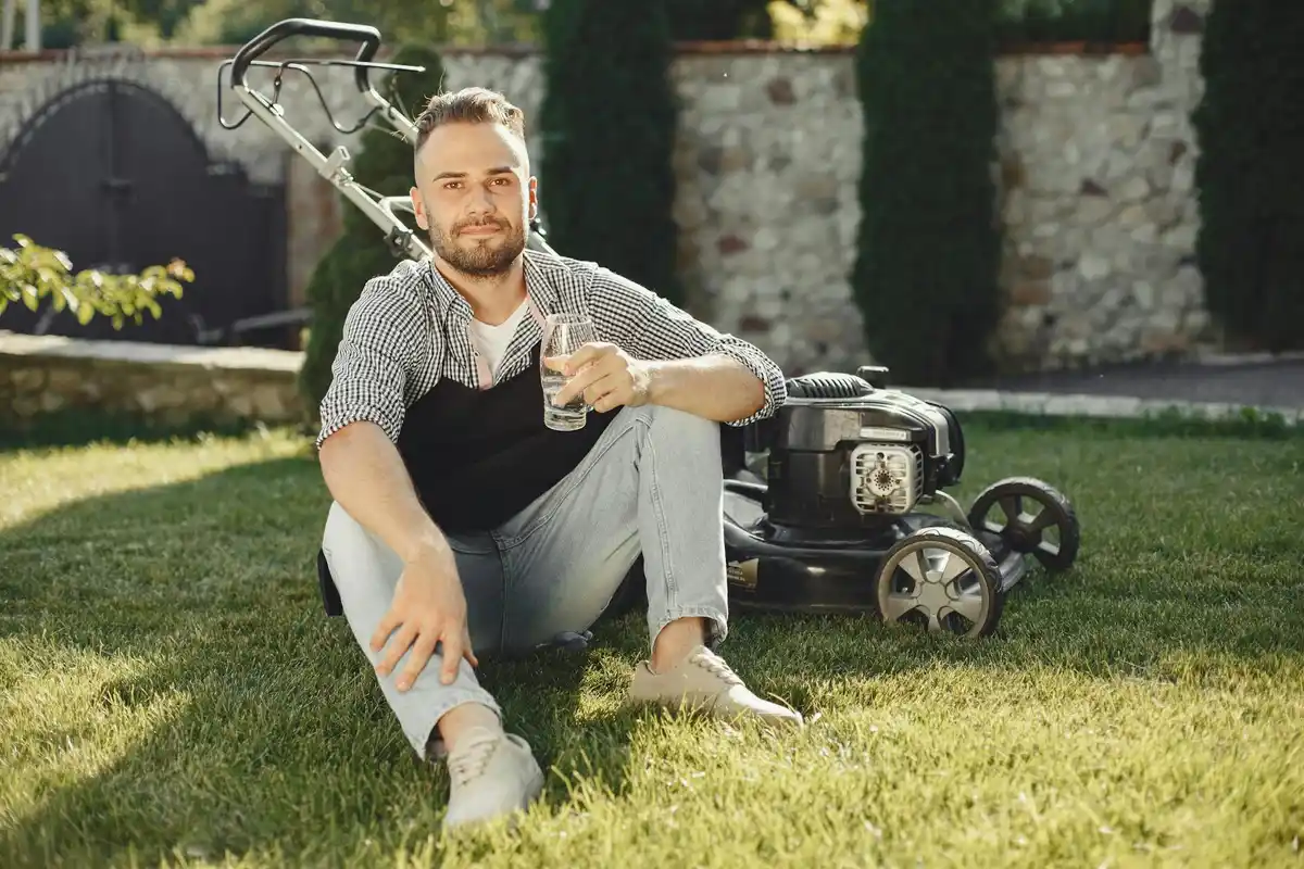 A man sits on a healthy green lawn with a lawn mower in the background, taking a break during yard work.
