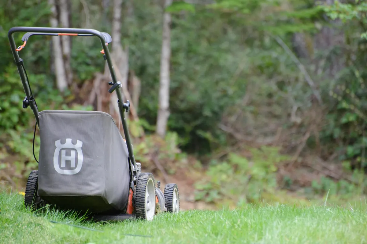 A lawn roller with a handle and grey cover sits on a green lawn, with trees and a garden in the background.