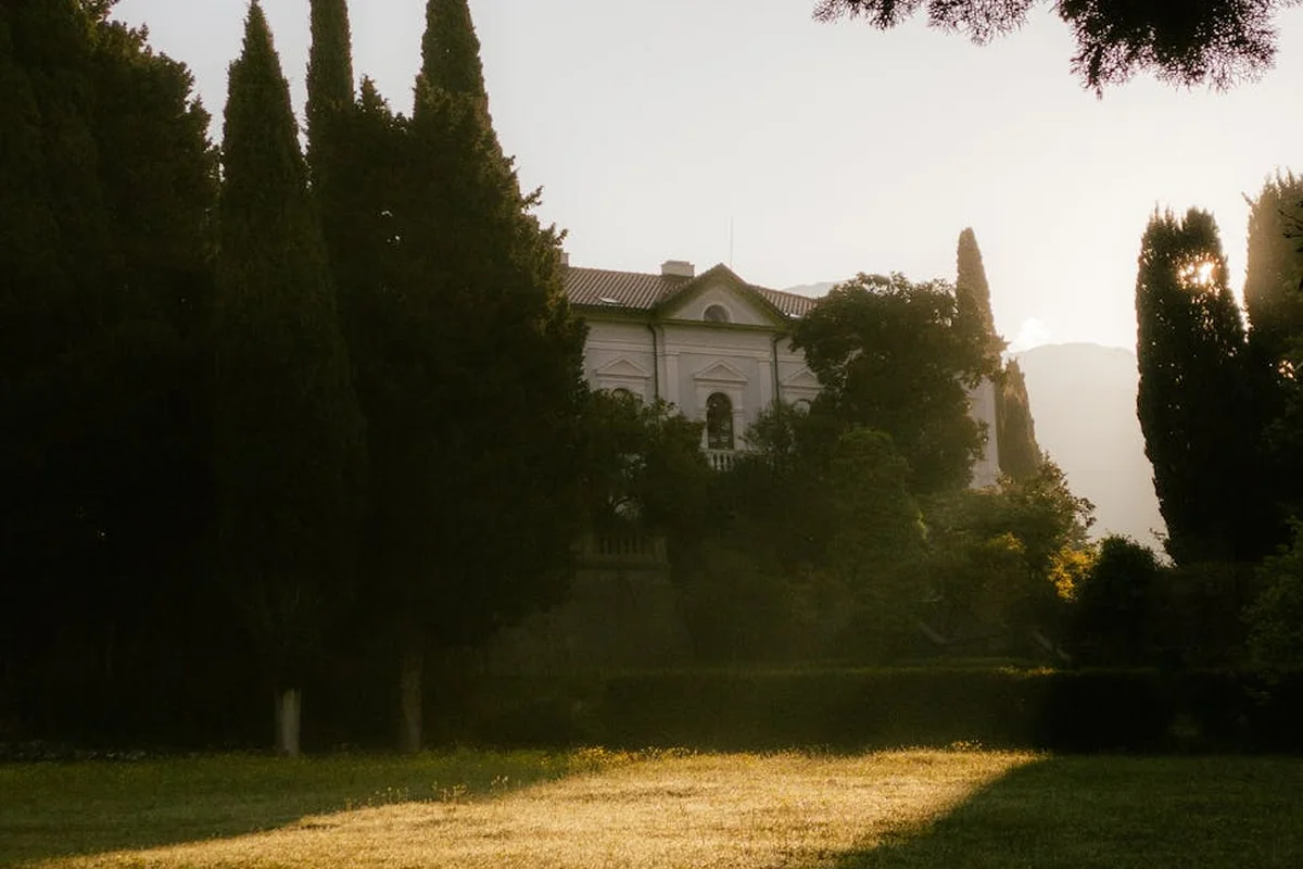 A wide, sunlit lawn in front of a stately house with tall trees casting long shadows.