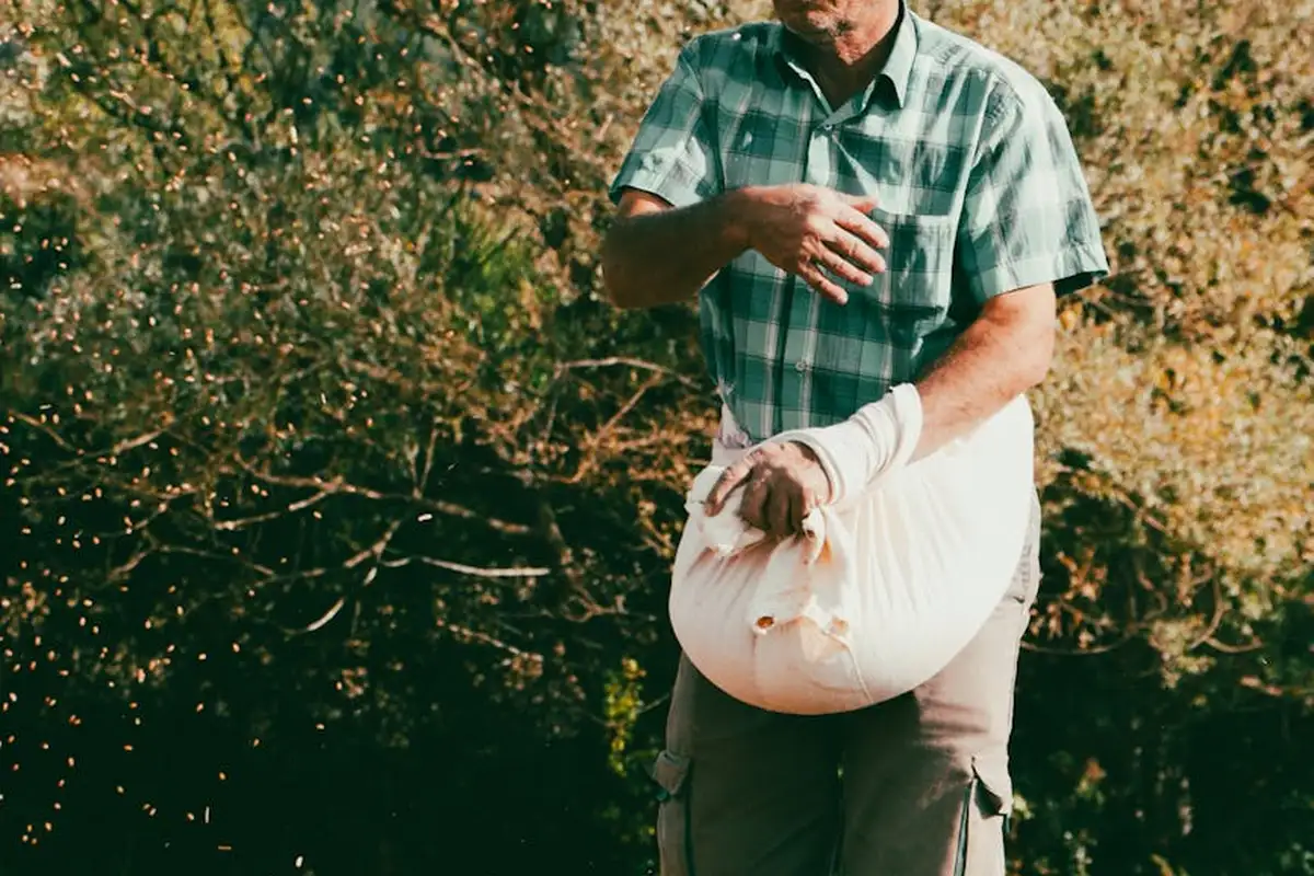 Man in a green plaid shirt outdoors holding a large white bag of lawn product, ready to seed or fertilize a lawn.