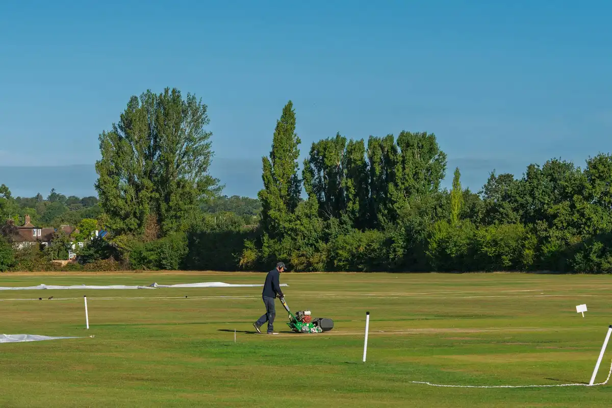 Groundskeeper mowing a large, flat lawn to create crisp mowing patterns with marker posts visible in the background.