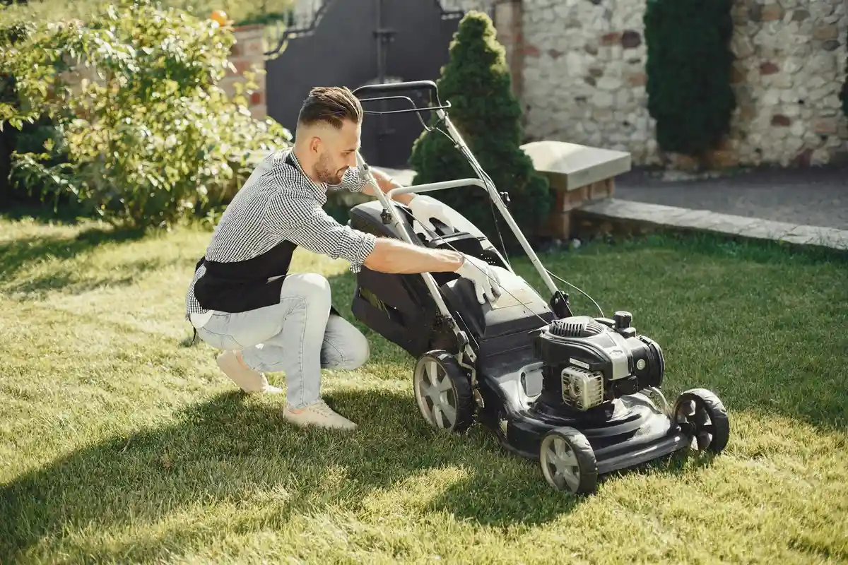 A man kneels on a well-kept lawn beside a push lawn mower, preparing to mow to create straight stripes.