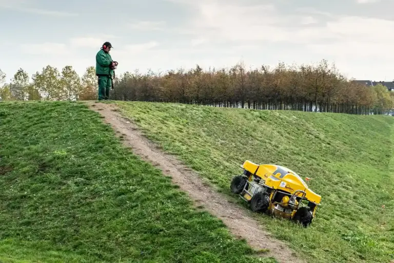Person in a green jacket standing on a grassy hill, operating a yellow fertilizer applicator on a slope