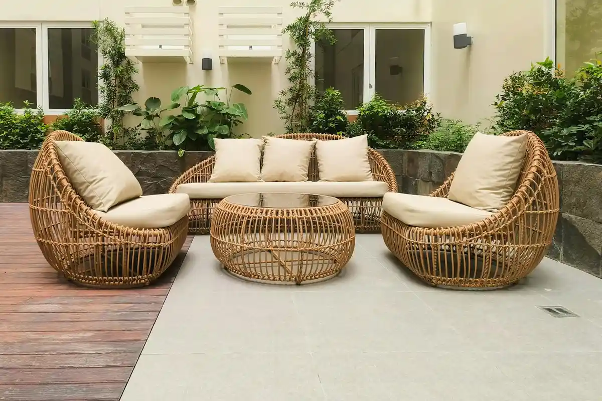 Outdoor wicker patio furniture with beige cushions arranged on a patio, with greenery and plants in the background.