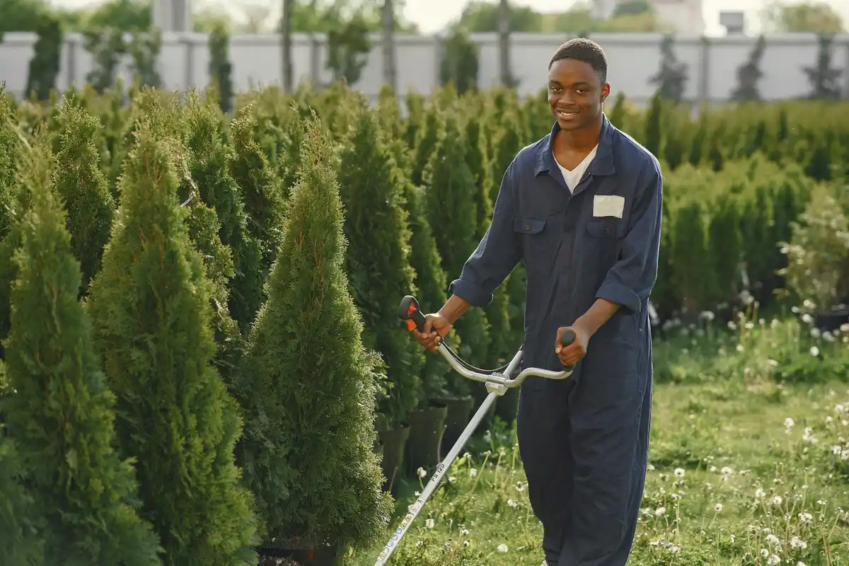 A technician in blue coveralls stands outdoors among tall evergreen shrubs, holding a long-handled yard tool.