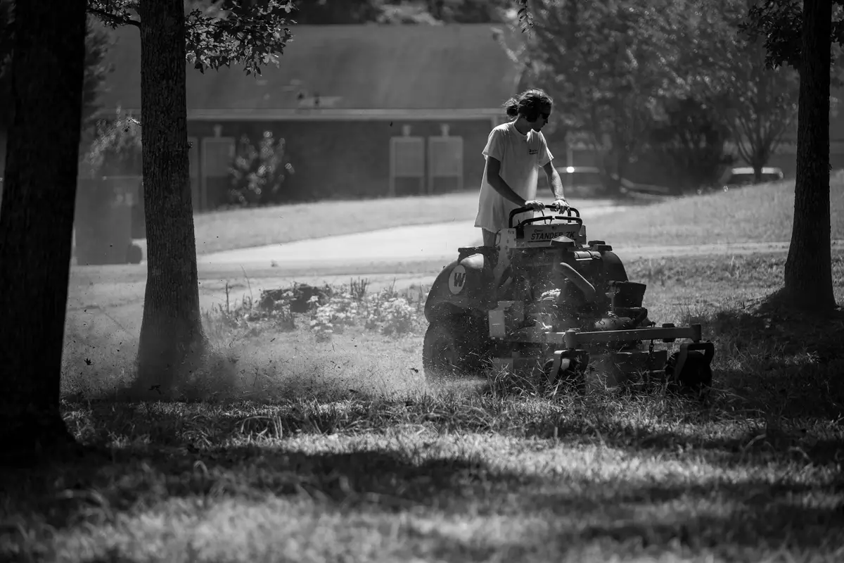 A person riding a lawn mower on a sunlit lawn surrounded by trees; black-and-white photograph.
