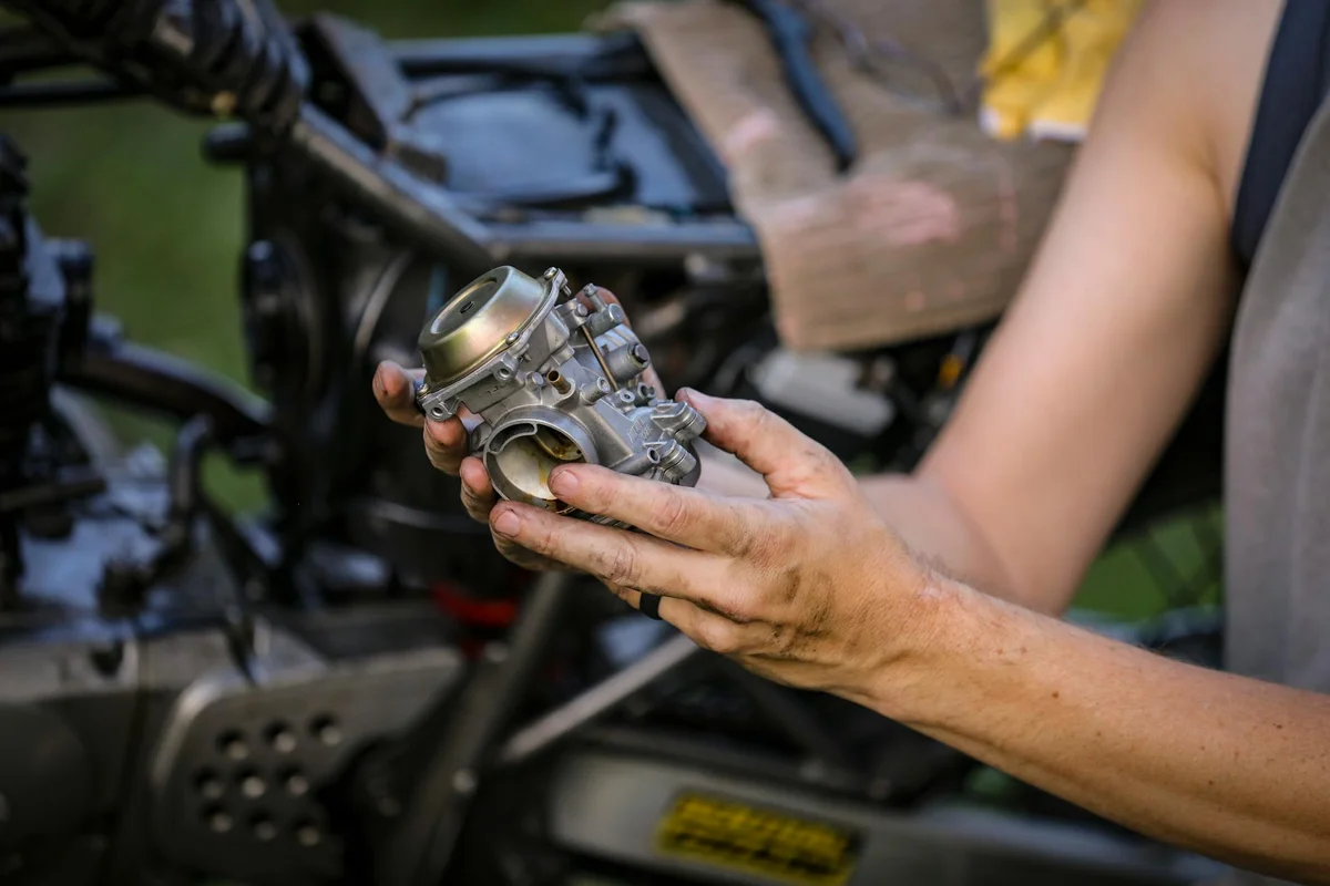Close-up of a person's hands holding a carburetor with a lawn mower blurred in the background.