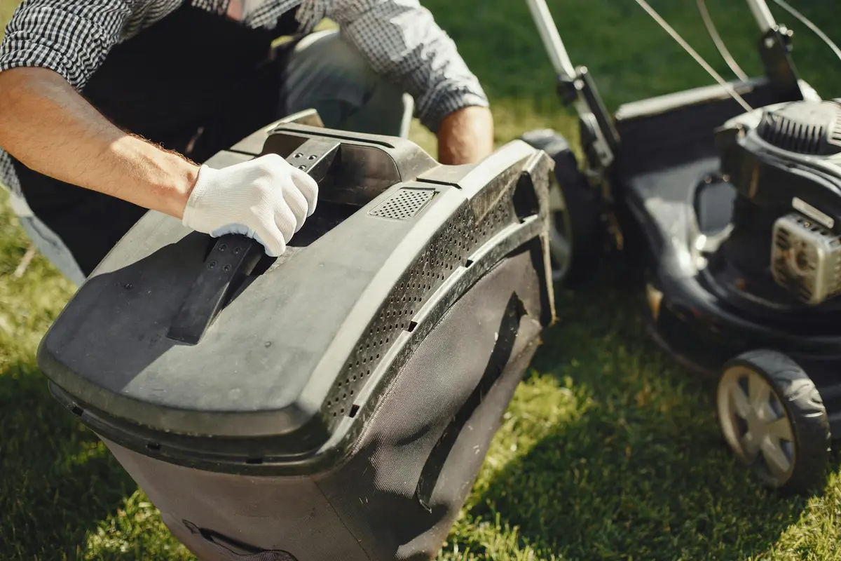 A person wearing gloves inspects the inner area of a lawn mower, lifting the cover near the engine to assess whether cleaning, repair, or replacement is needed.