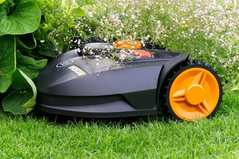 A robotic lawn mower on green grass with an orange wheel beside leafy plants.