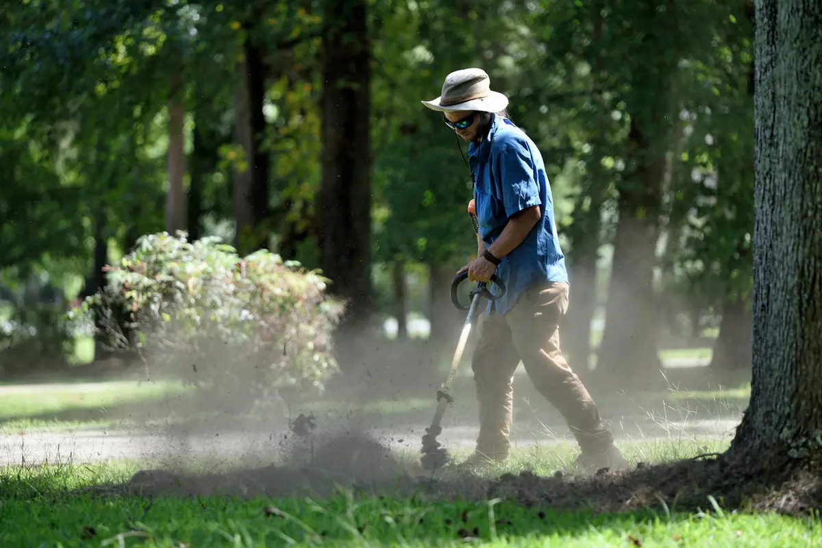 Person wearing a wide-brim hat and sunglasses mowing a grassy lawn among trees.