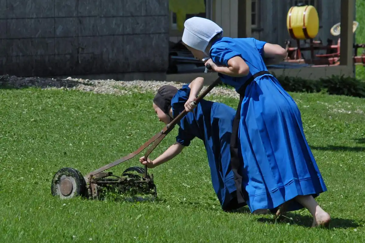Two people in blue outfits inspecting a lawn mower on a grassy lawn.