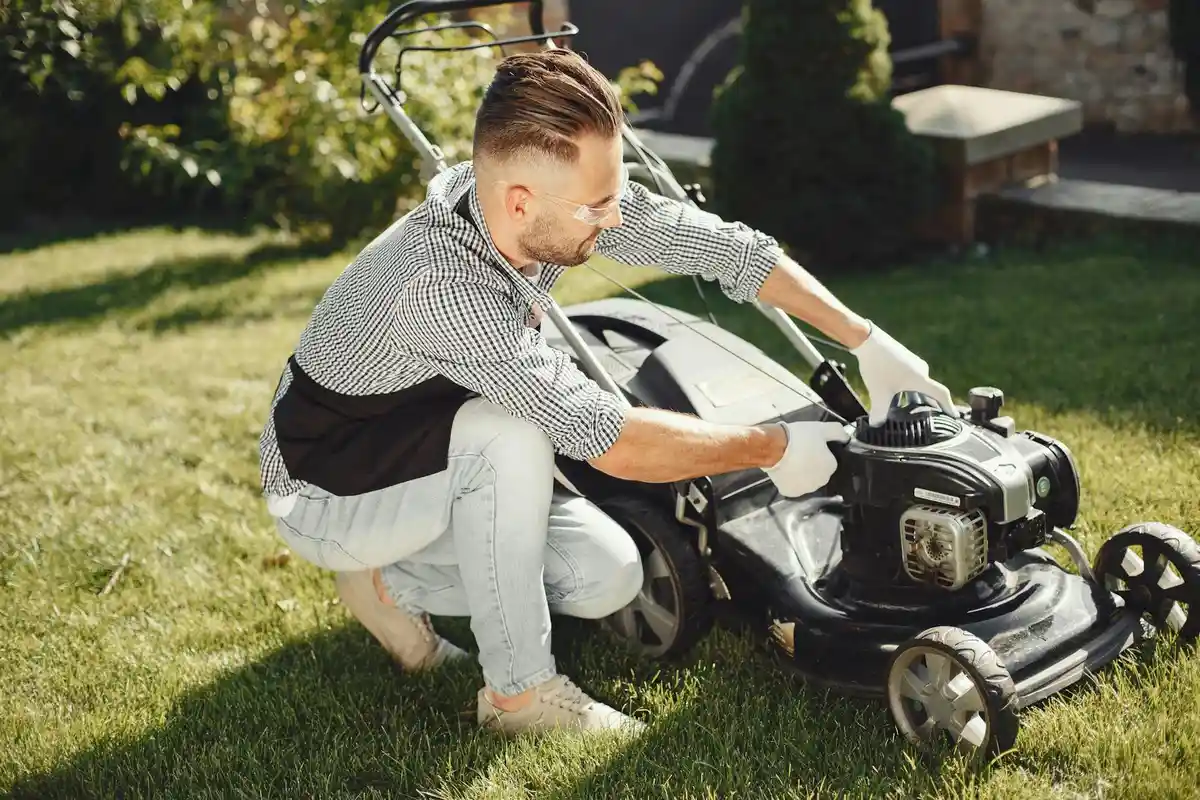 A person crouches beside a gas-powered lawn mower on a green lawn, inspecting the mower before mowing.