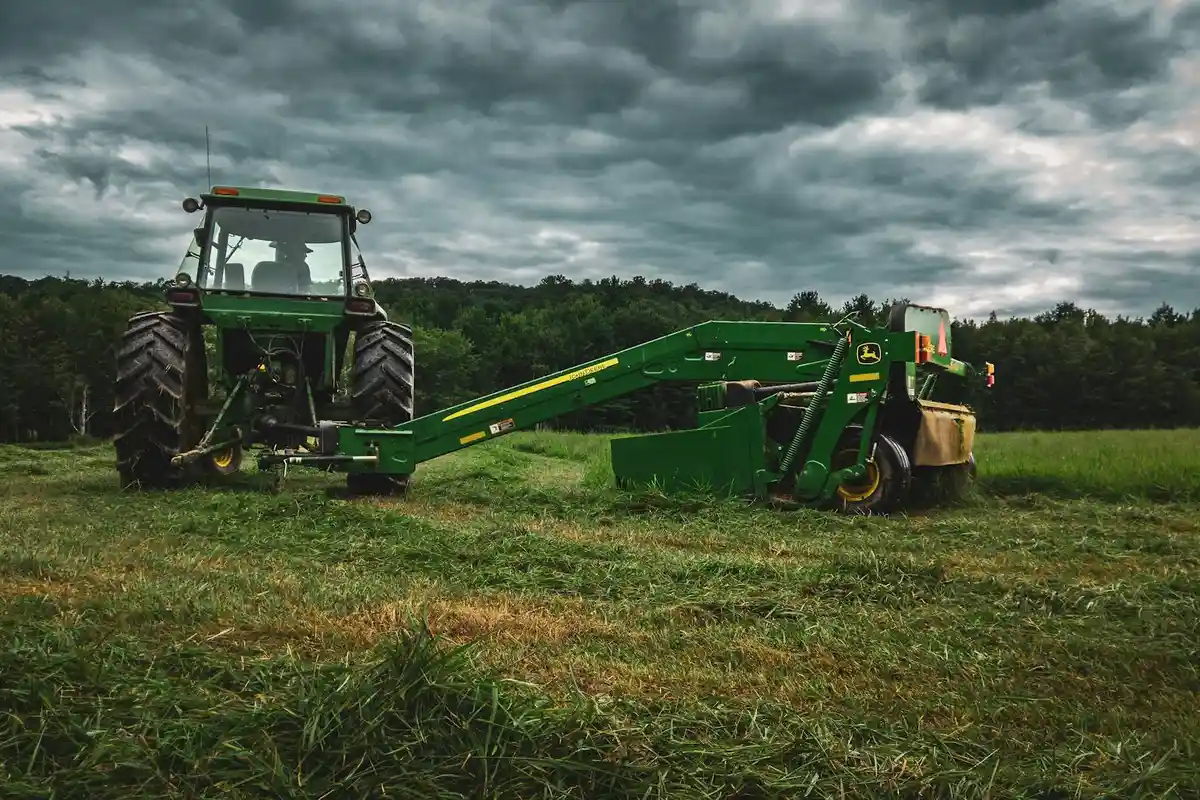 Green tractor with a mower attachment in a grassy field under a cloudy sky